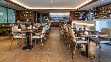 Ein modernes Restaurant mit Holztischen und -stühlen, einer Bar mit Regalen voller Flaschen und Blick auf Berge durch das Fenster.