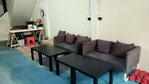 A room with a grey sofa, two black tables, a red stool, and a white desk with a clock and some items on it. - First Inn On Nut - Hostel (Bangkok)