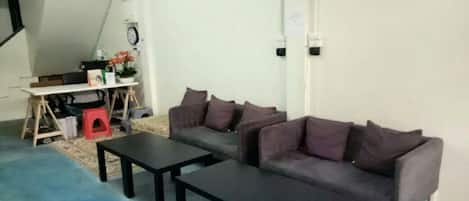 A room with a grey sofa, two black tables, a red stool, and a white desk with a clock and some items on it.