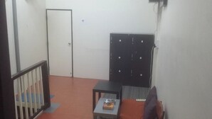 A small room with a door, a black cabinet, a small table with a book, and a brown chair. - First Inn On Nut - Hostel (Bangkok)