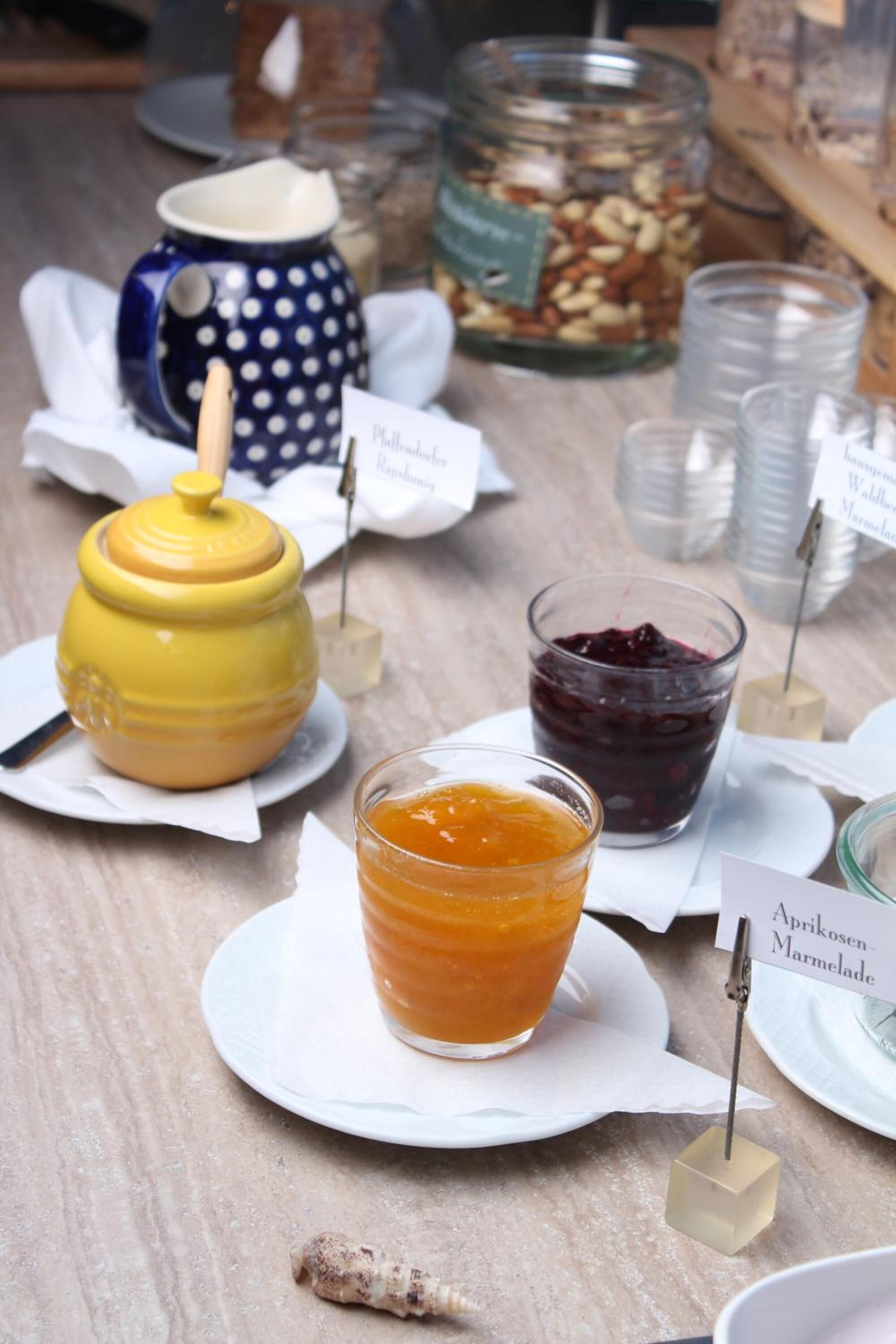 Ein Tisch mit einem Honigglas, einem Glas Honig, einem Aprikosenmarmeladenglas und einer gepunkteten Tasse.