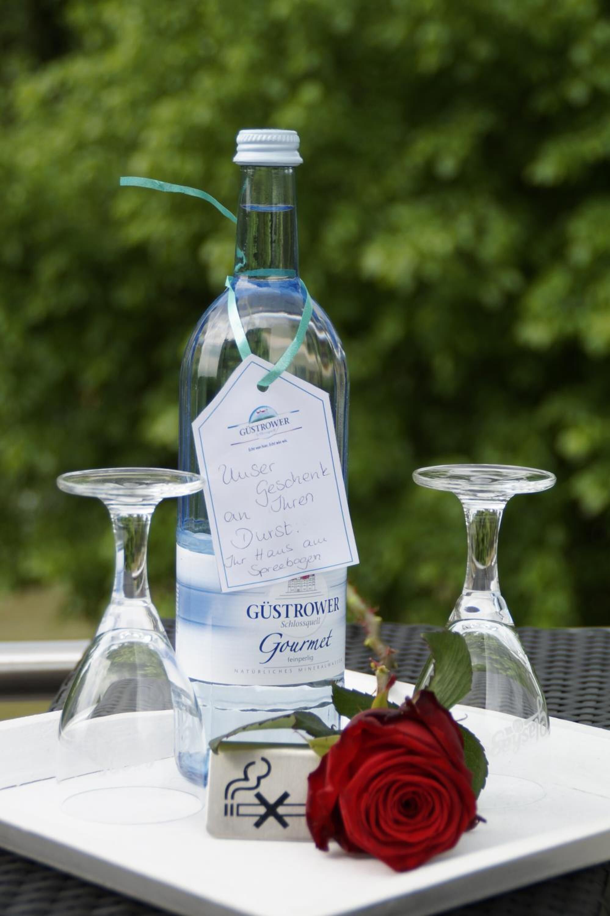 A bottle of Gustrower Gourmet wine with a tag, two clear wine glasses, and a red rose on a white tray.