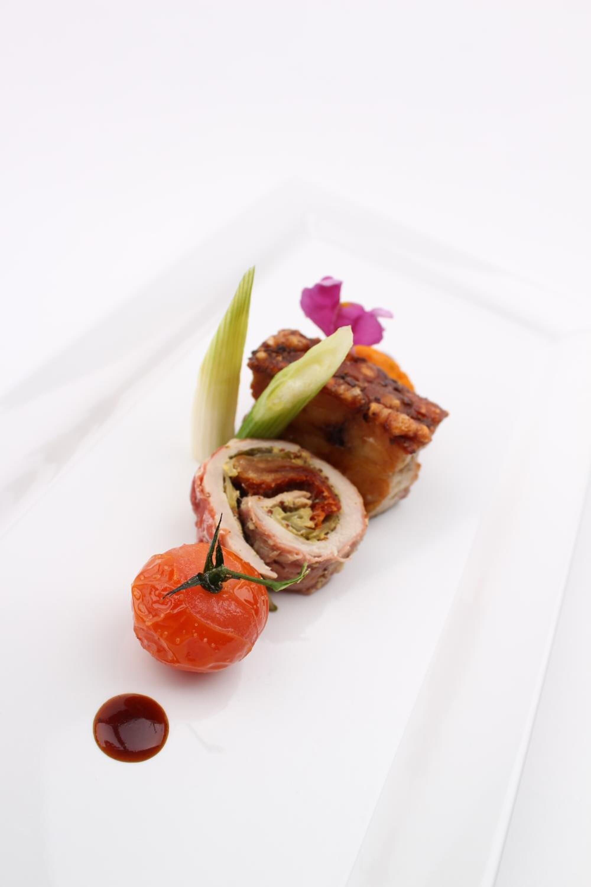 A white plate with a rolled meat dish, a cherry tomato, and a small flower.