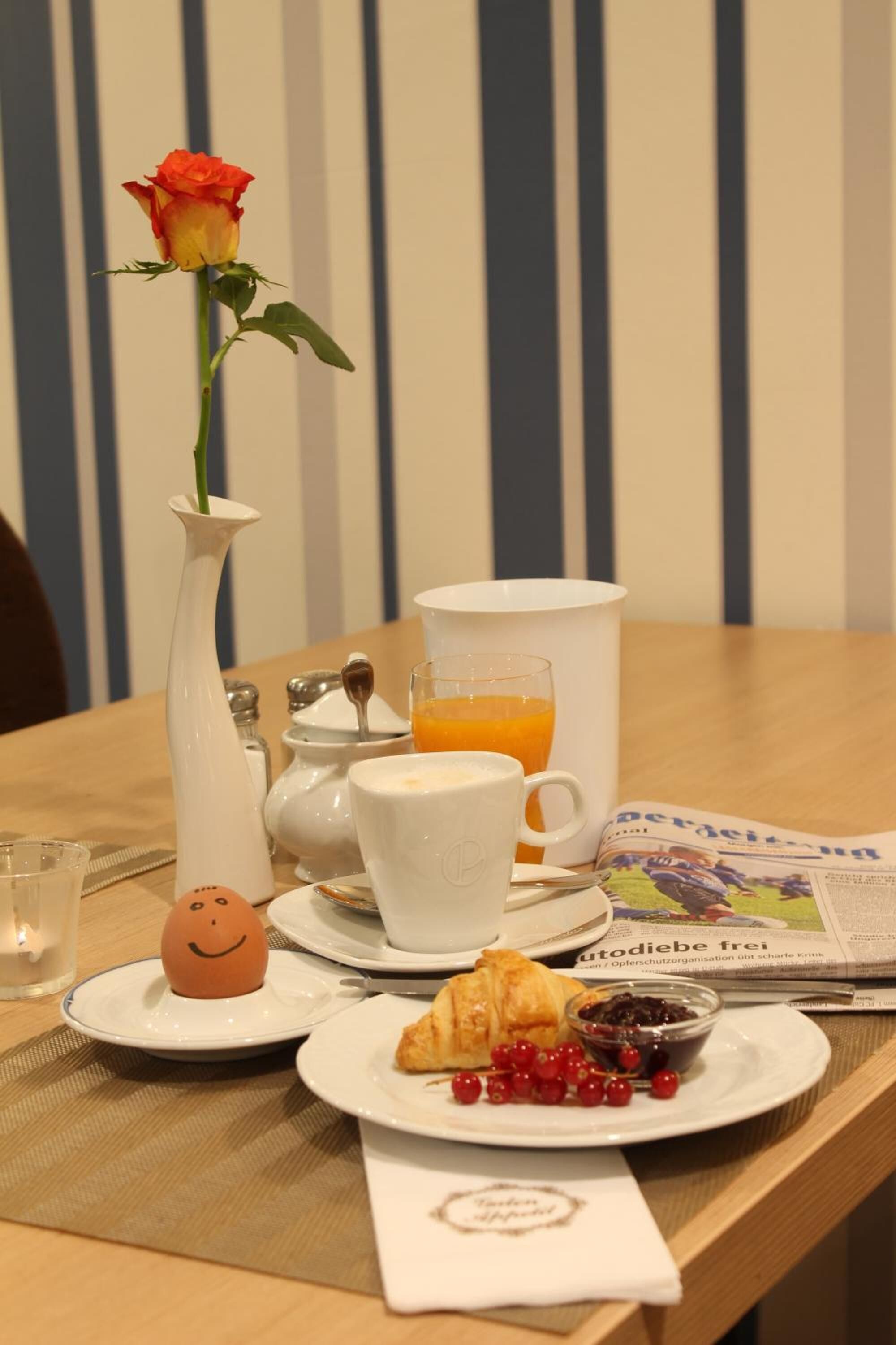 Een ontbijttafel met een vaas met één roos, een kop koffie, een glas sinaasappelsap, een croissant en een bord met bessen.