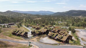 A cluster of traditional wooden cabins with grass roofs, surrounded by a forested landscape and mountains in the background.