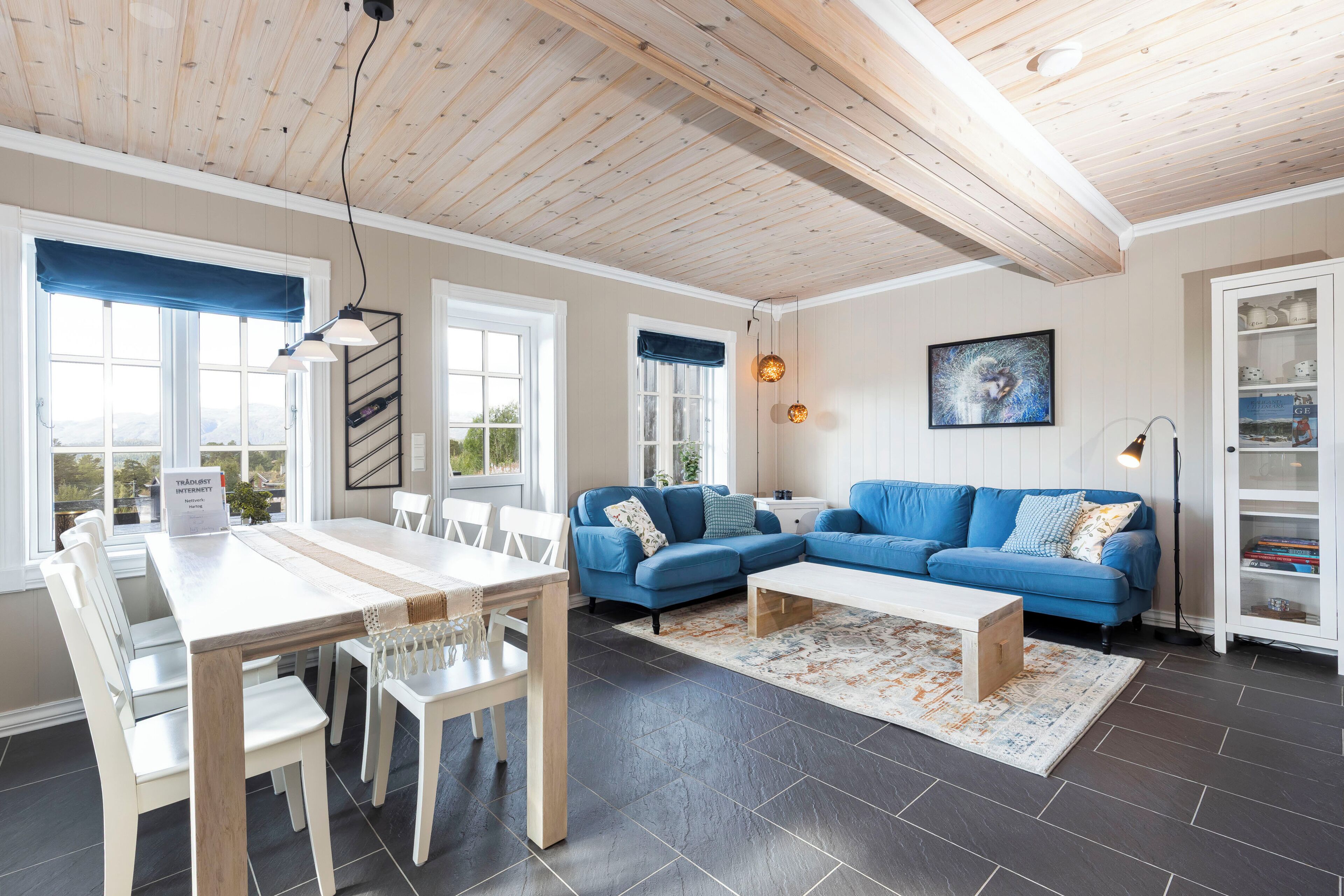 Uma sala de estar moderna com área de jantar, sofás azuis, mesa branca e teto de madeira.