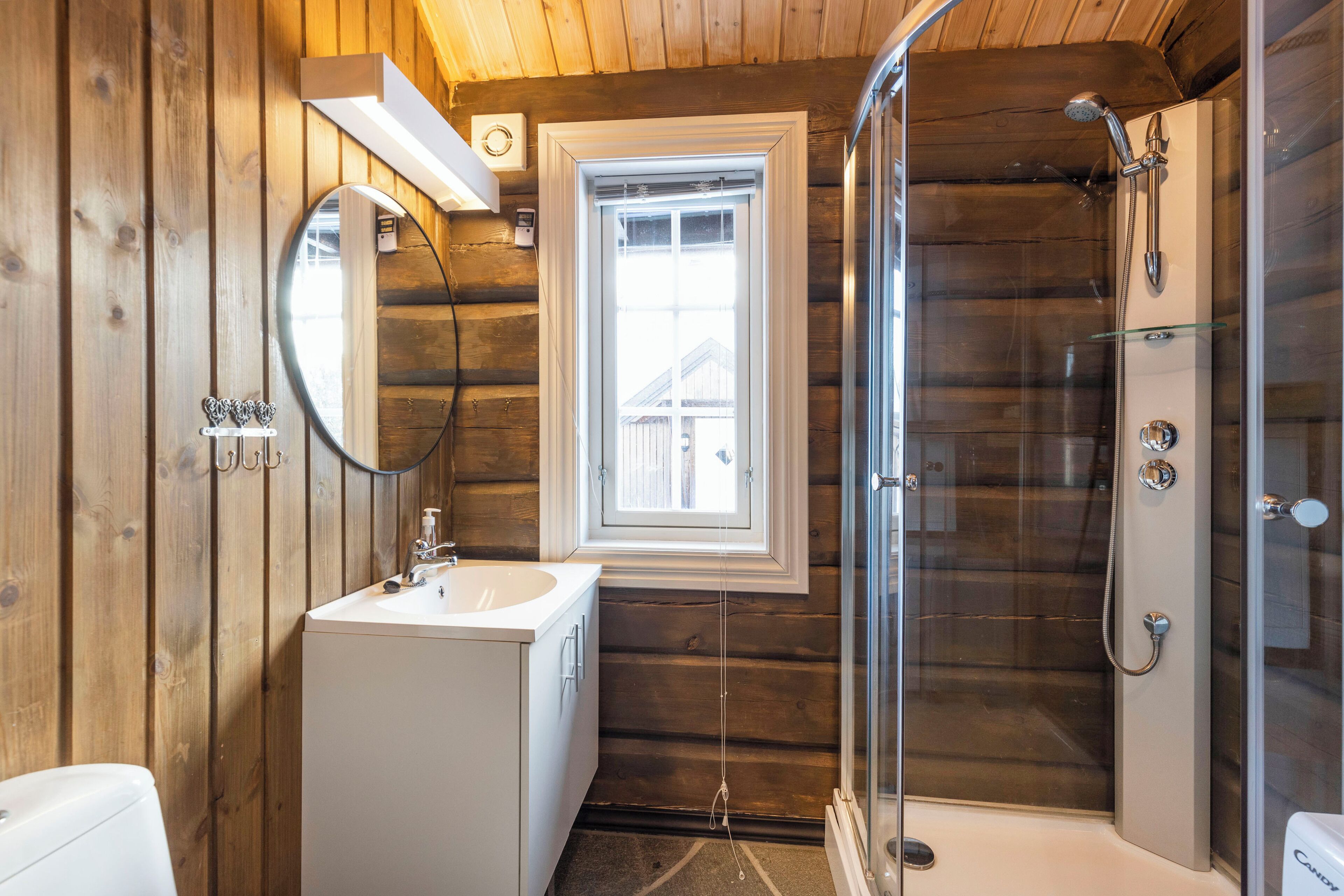 Een houten badkamer in een vakantiehuisje, met een wastafel, spiegel, raam en douche.