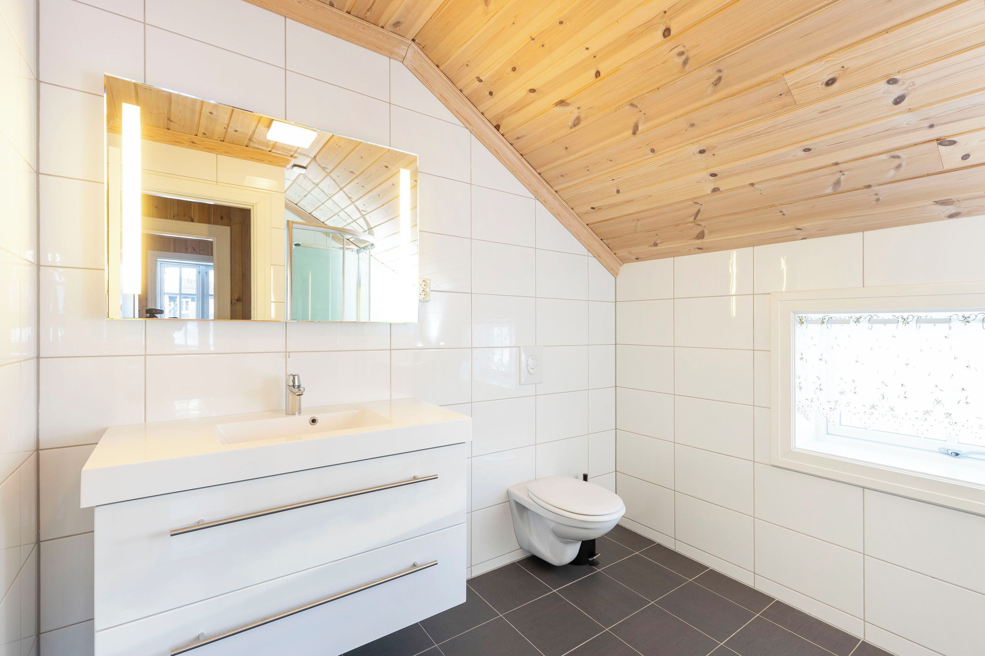 A modern bathroom with a wooden ceiling, white tiles, a sink with a mirror, and a toilet.