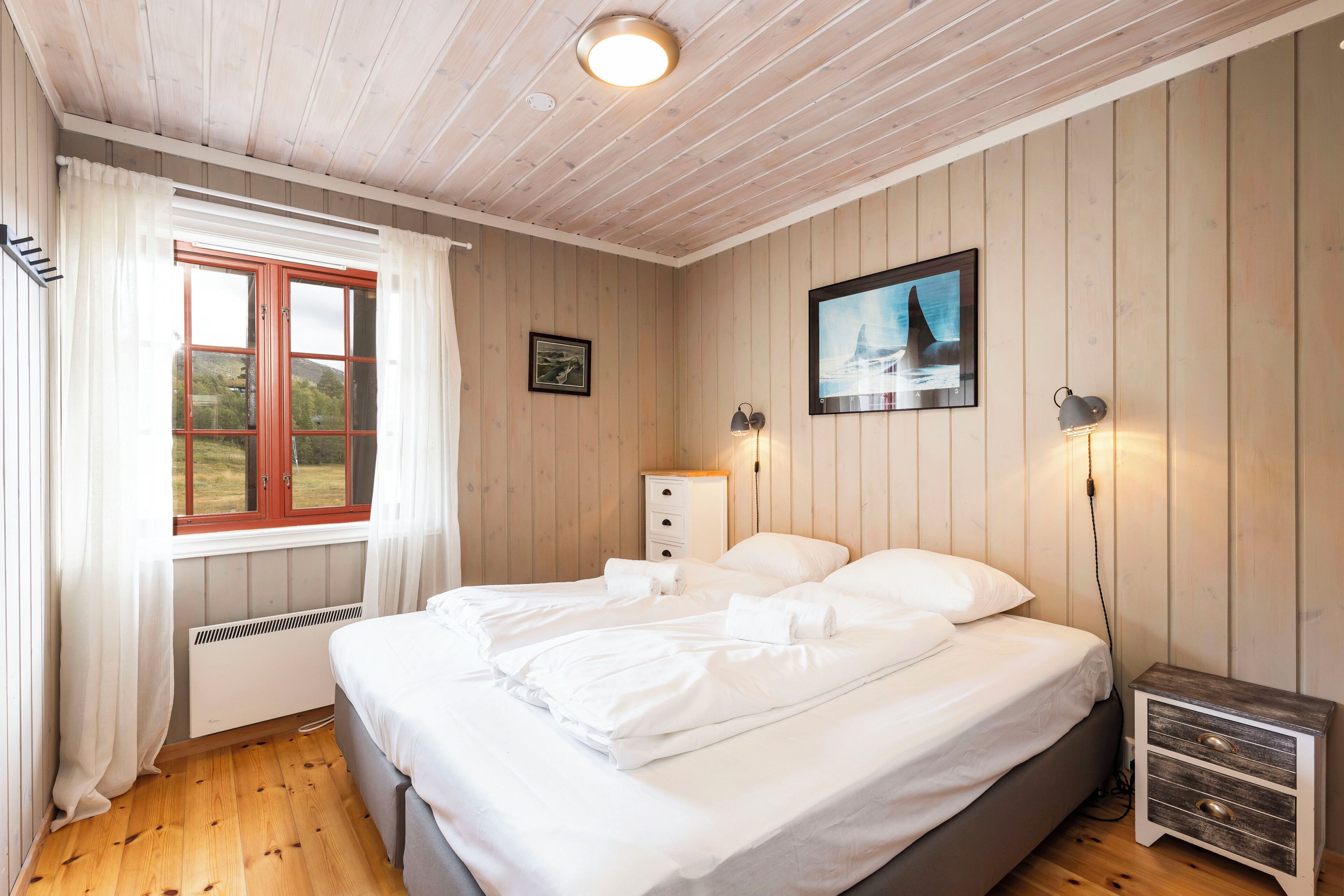 A bedroom with a bed, a nightstand, a lamp, a window with curtains, and a wall-mounted picture.