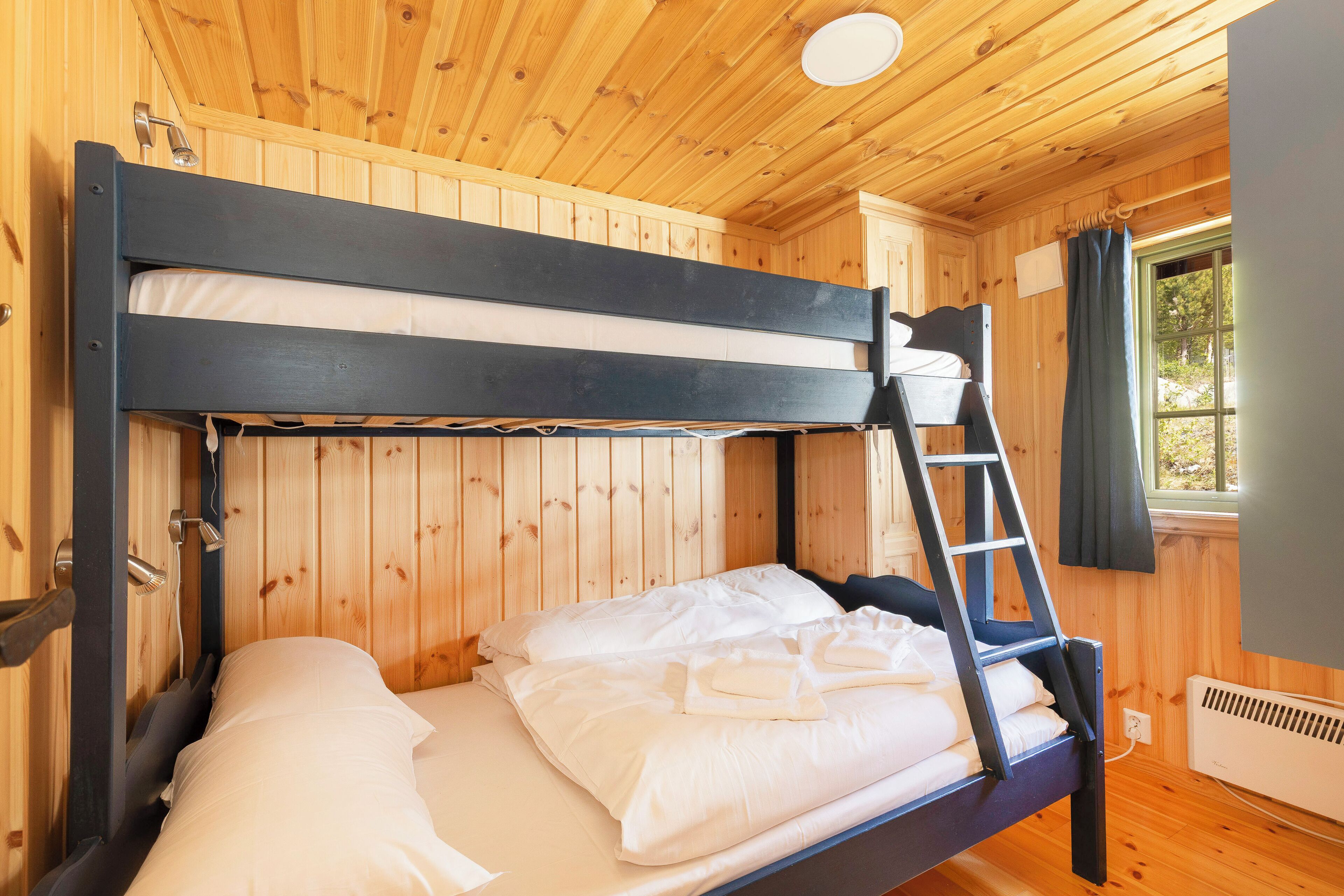 Uma cama de beliche com escada, paredes de madeira, uma janela com cortinas e um radiador.