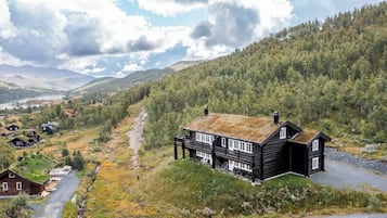 Uma casa de madeira com telhado de palha, rodeada por uma encosta arborizada e um lago ao fundo.