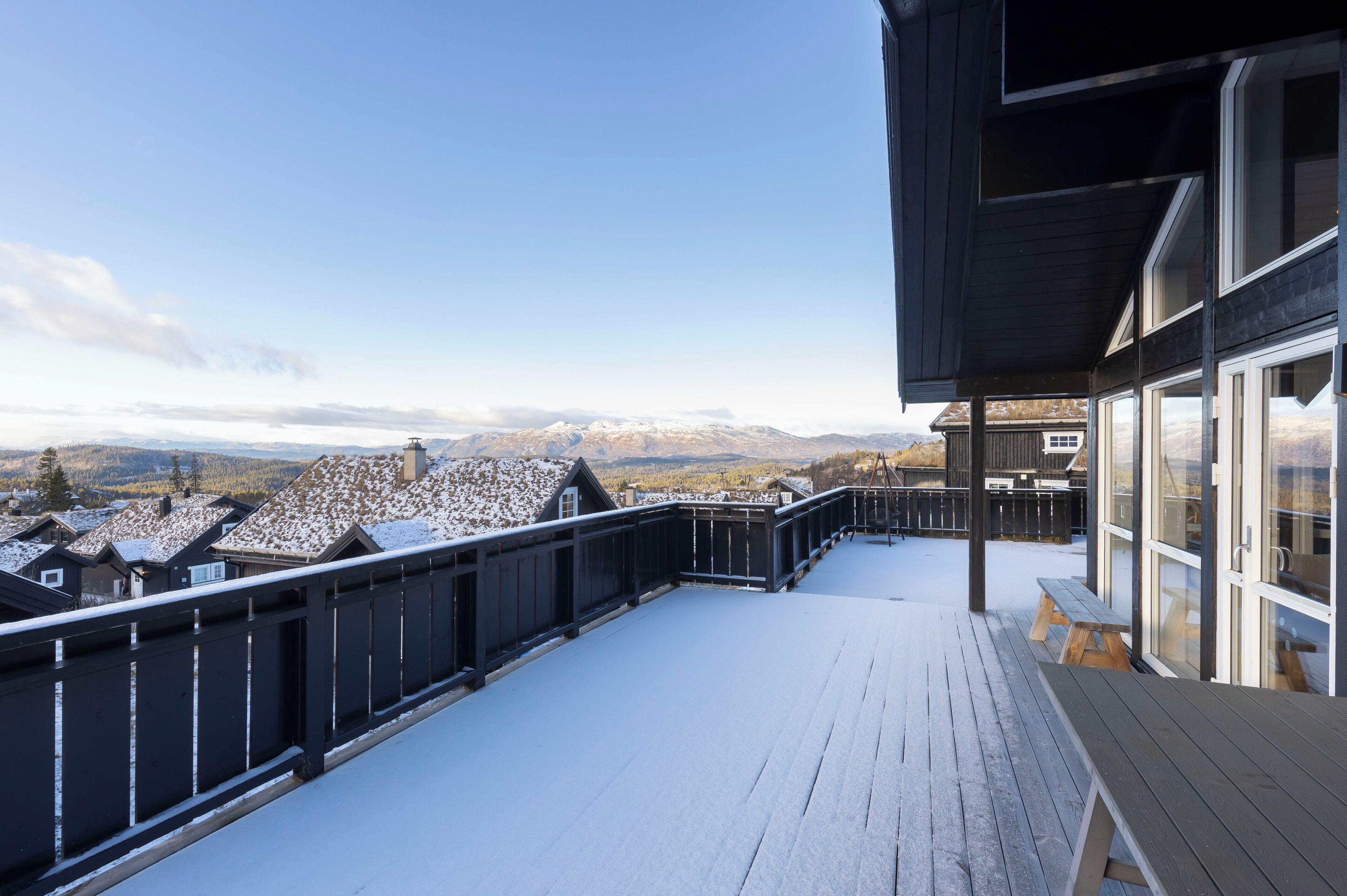 A wooden deck with a railing, overlooking a snowy landscape with houses and mountains.