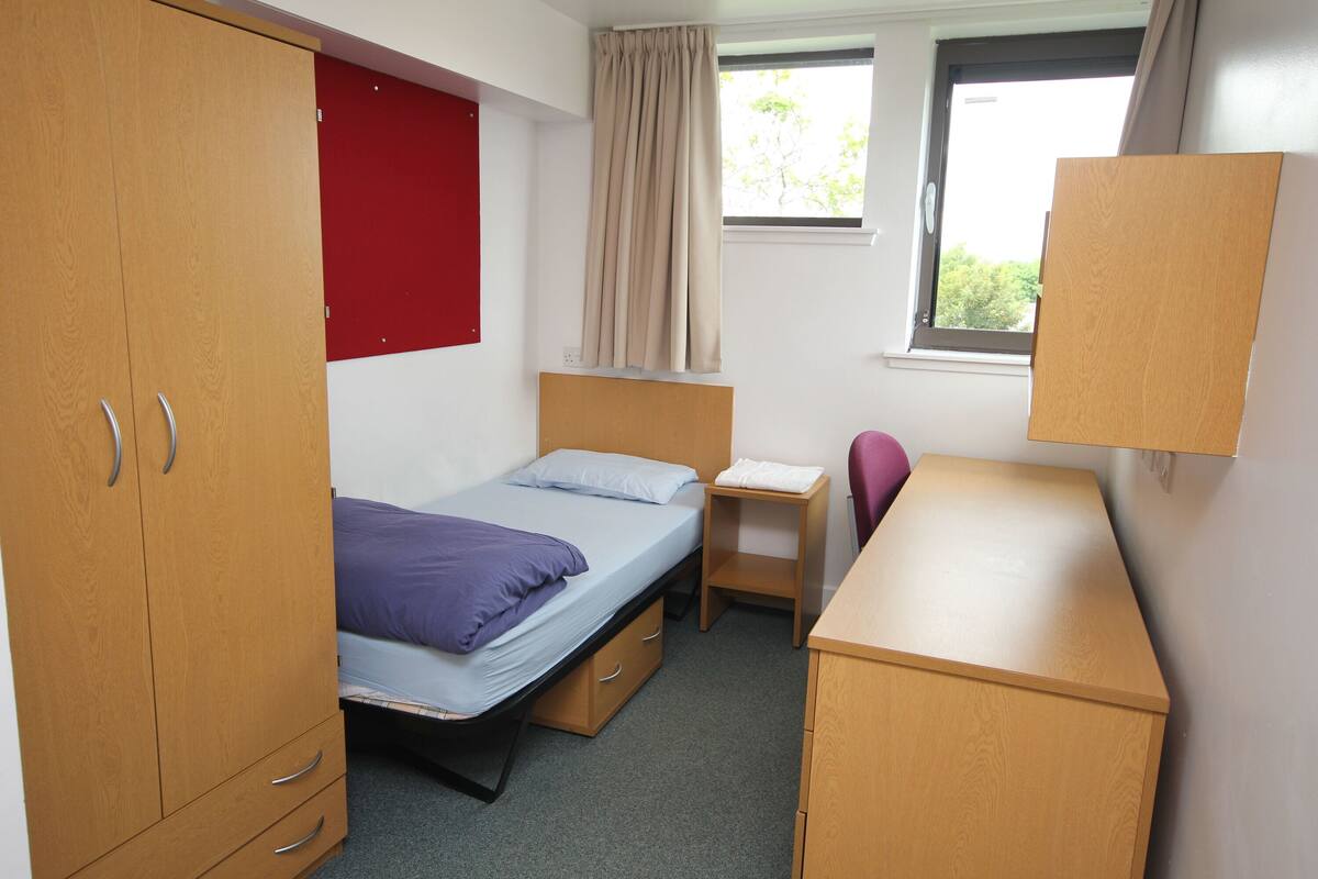 A dormitory room with a bed, desk, chair, wardrobe, and a window with curtains.