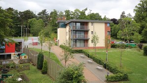 Un immeuble moderne à plusieurs étages, doté de balcons, entouré de verdure et bordé d’un chemin pavé.