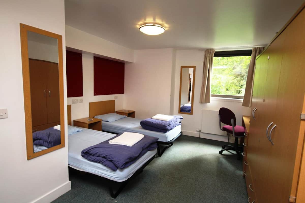 A dormitory room with two single beds, a wooden wardrobe, a mirror, a desk, and a chair.