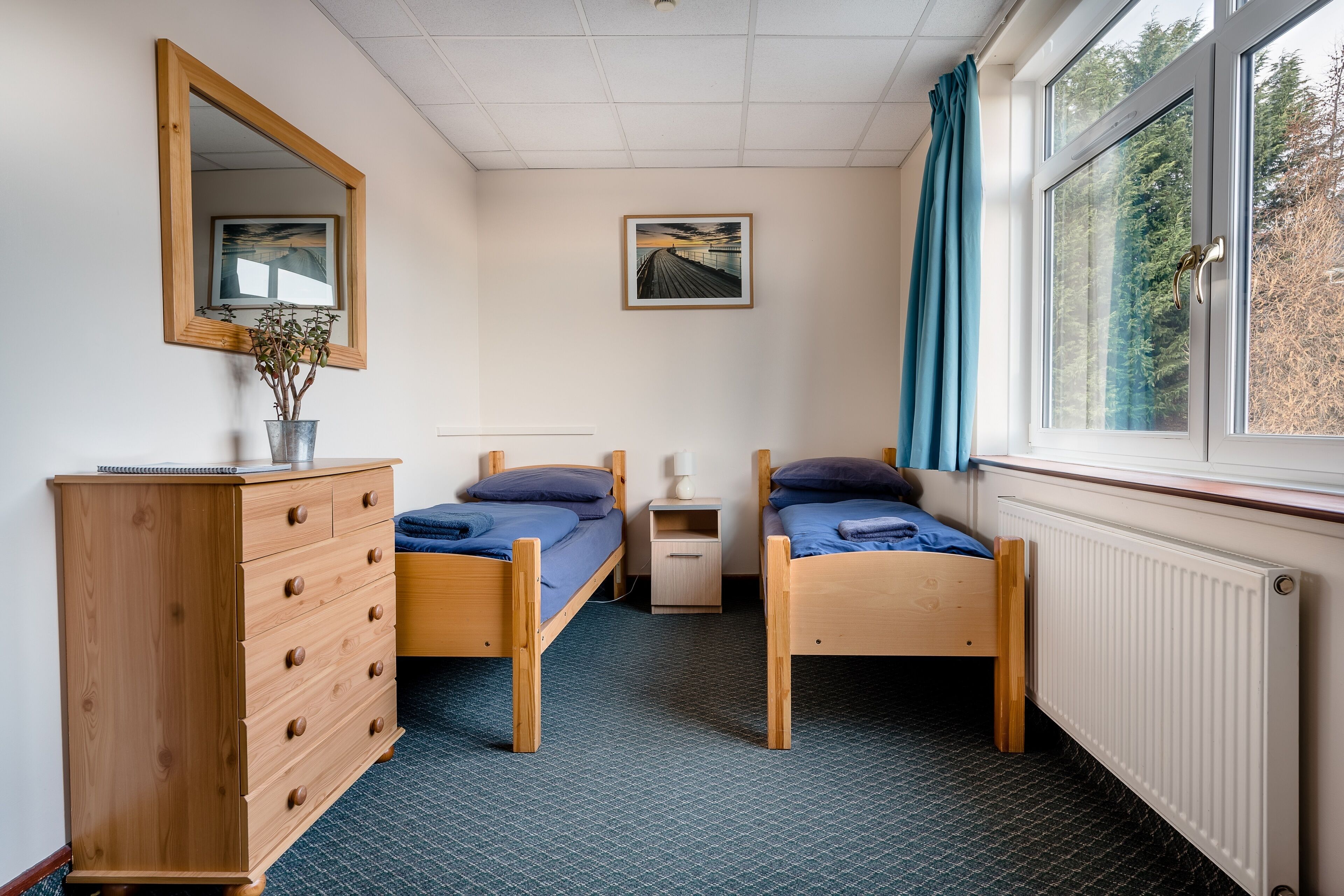A bedroom with two single beds, a wooden dresser, a mirror, a framed picture on the wall, and a window with curtains.
