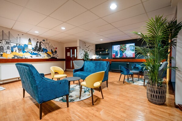 A lobby with blue and yellow chairs, a wooden floor, a large mural, and a television screen.