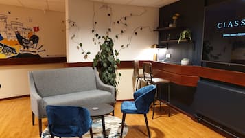 Uma área de estar acolhedora, com um sofá cinzento, duas poltronas azuis, uma mesinha redonda e uma planta decorativa. Uma estante com livros e uma luminária são visíveis ao fundo.