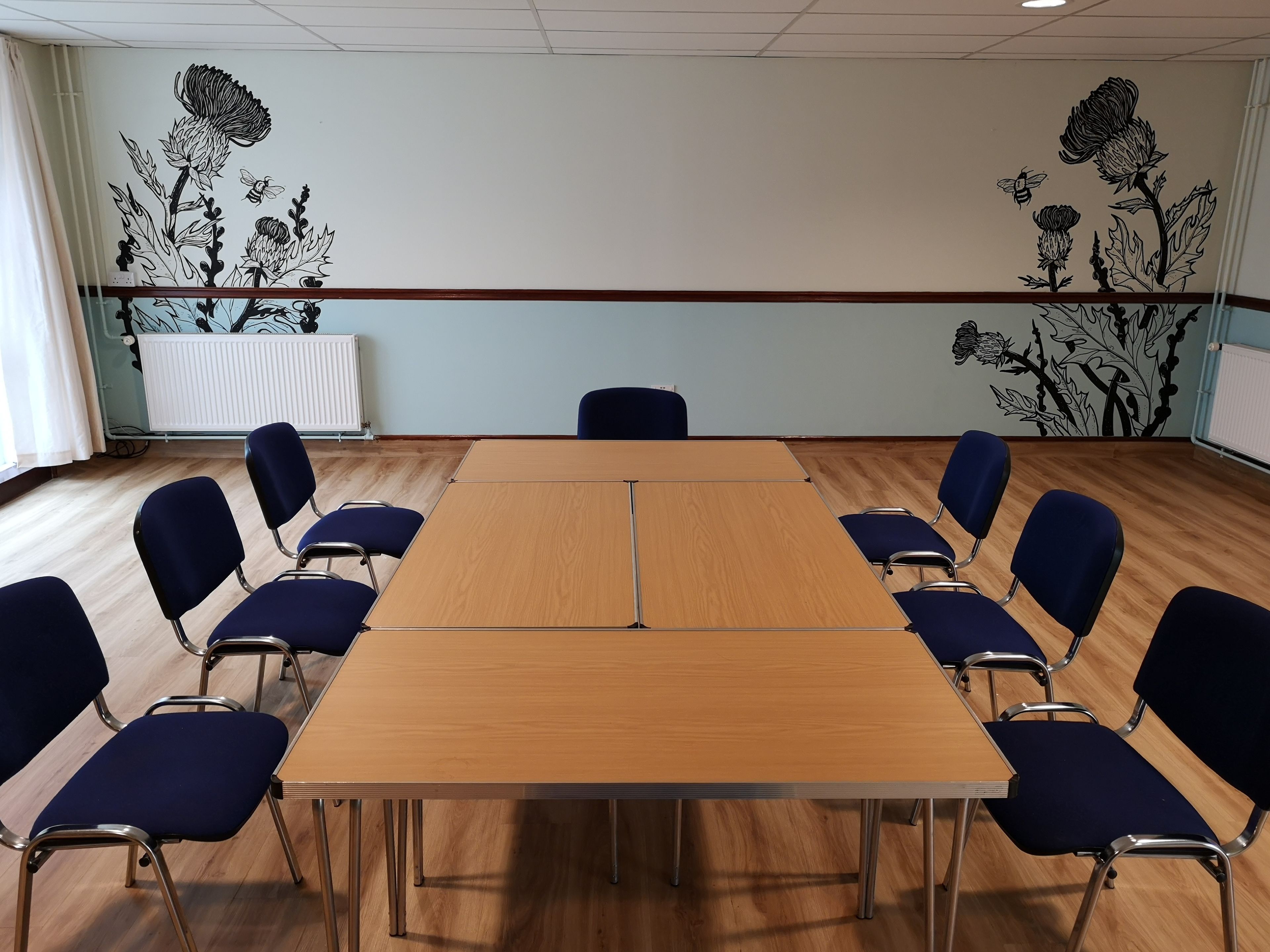 Un salón de conferencias con una larga mesa, sillas azules y decoración floral en la pared.