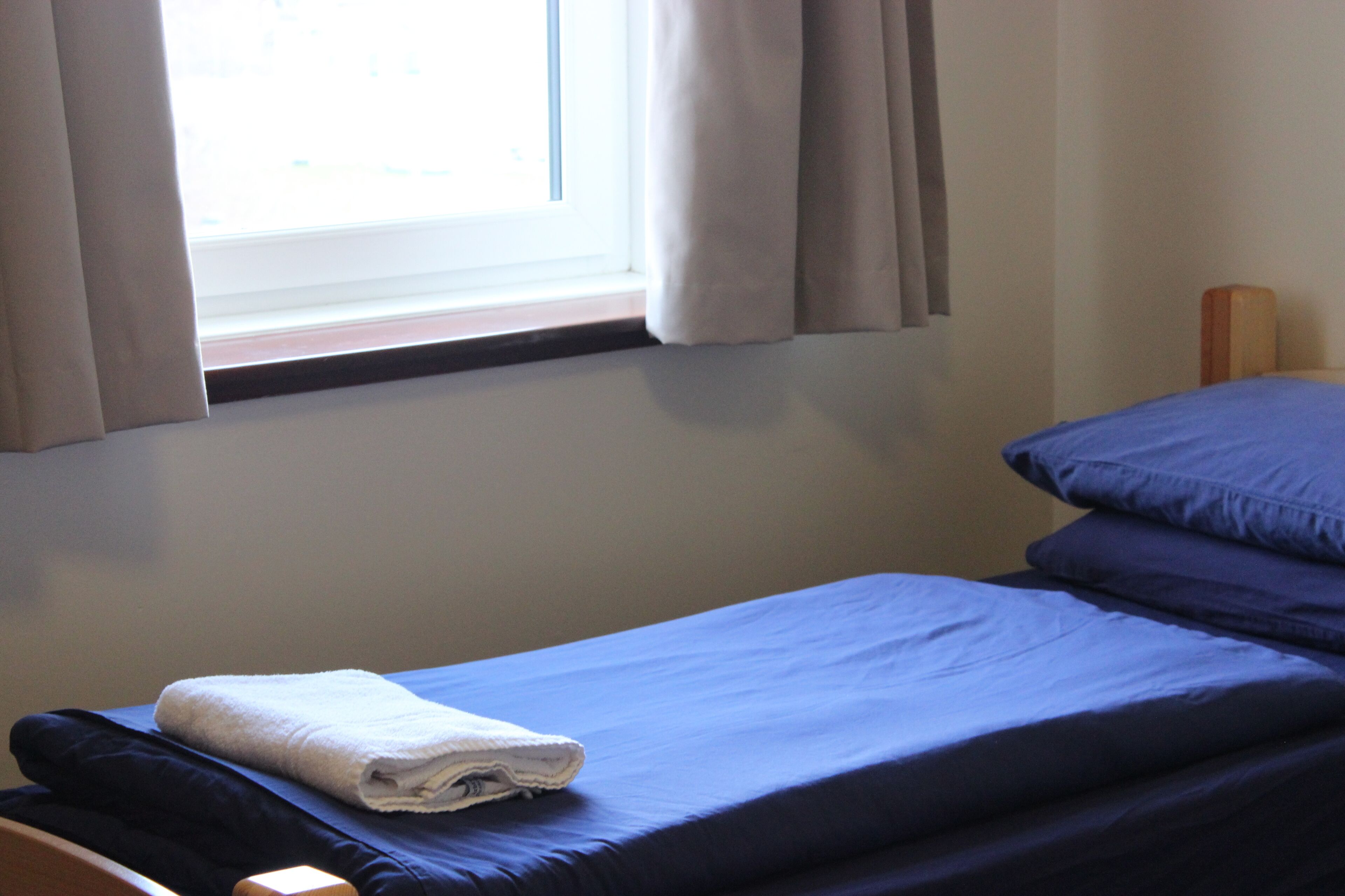 Una cama individual con colchón y almohadas azules, una toalla blanca y una ventana con cortinas.