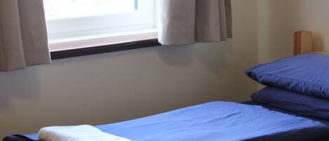 Una cama individual con colchón y almohadas azules, una toalla blanca y una ventana con cortinas.