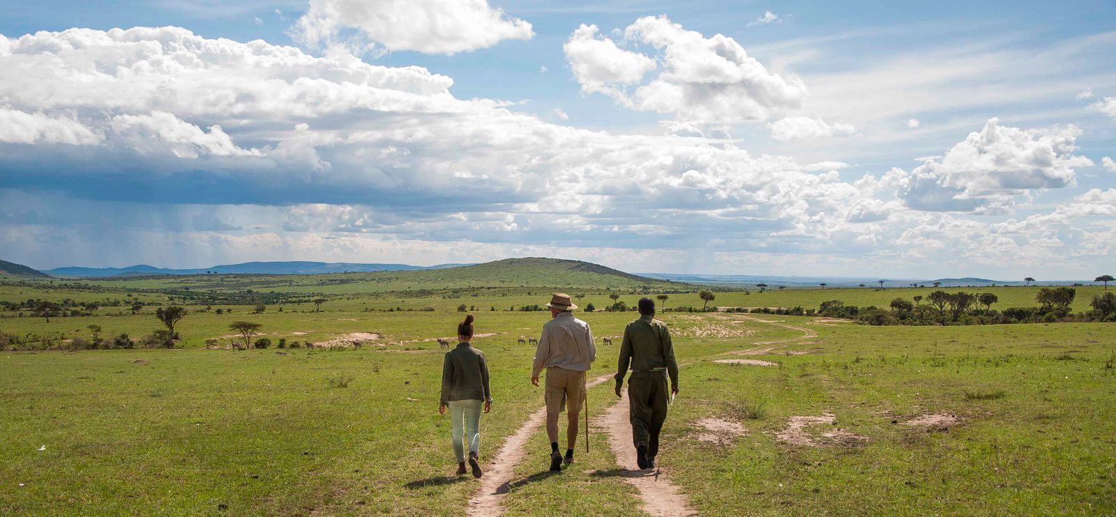 A group of people walking on a dirt path in a grassy field with distant hills and a partly cloudy sky.