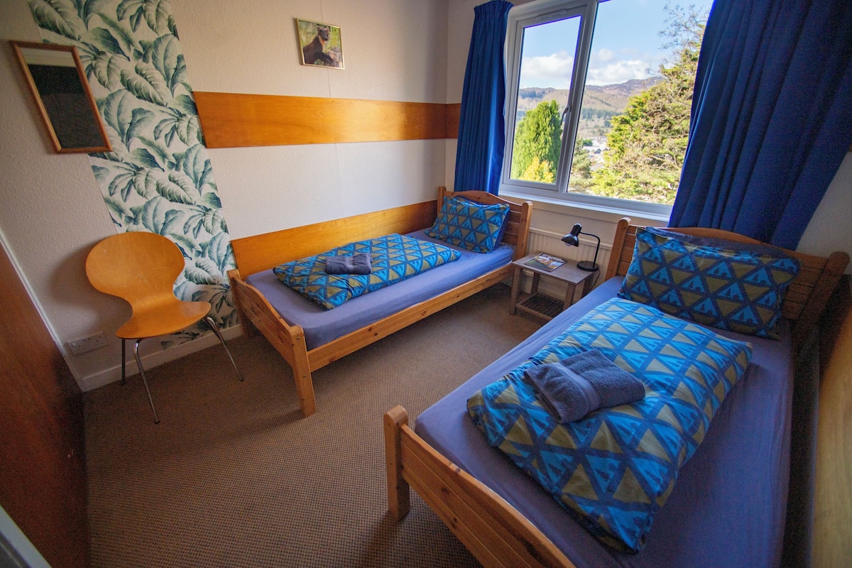 Quarto com duas camas de solteiro, uma cadeira de madeira, uma mesinha e janela com vista para áreas verdes e colinas.