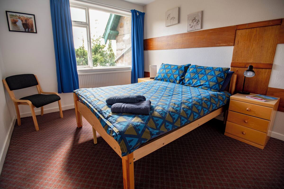 Quarto com cama, uma cadeira, mesa de cabeceira e janela com cortinas.