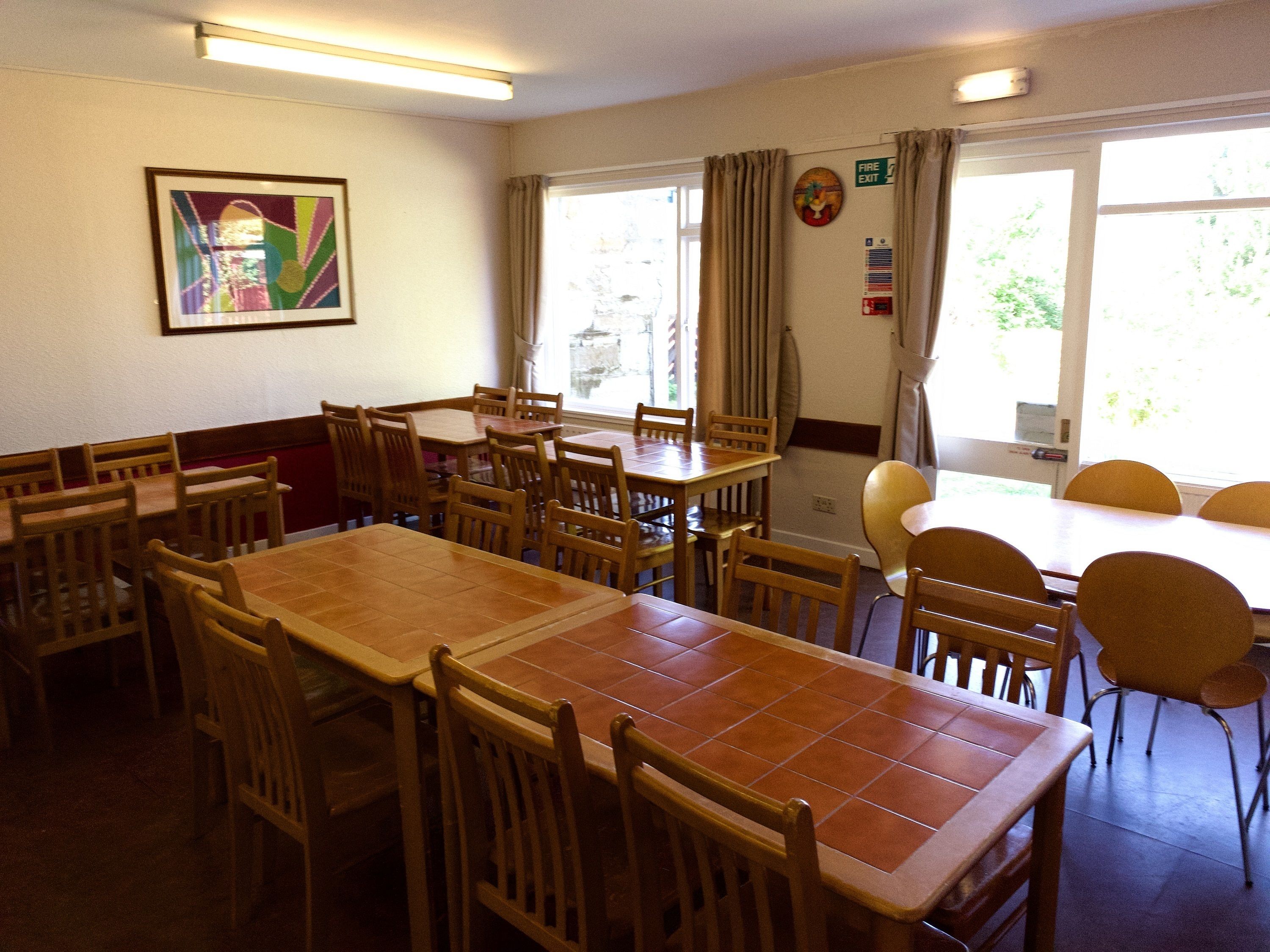 A dining area with multiple wooden tables and chairs, a framed artwork on the wall, and a window with curtains.