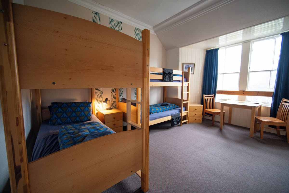 Quarto com beliche, escrivaninha e cadeira, janela com cortinas e estante.