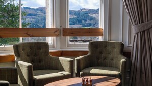 A room with two green upholstered chairs, a wooden table, and a view of a mountainous landscape through large windows. - Pitlochry Youth Hostel (Pitlochry)