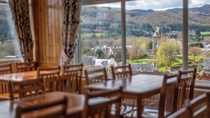 A dining area with wooden tables and chairs, large windows offering a view of a village and mountains. - Pitlochry Youth Hostel (Pitlochry)
