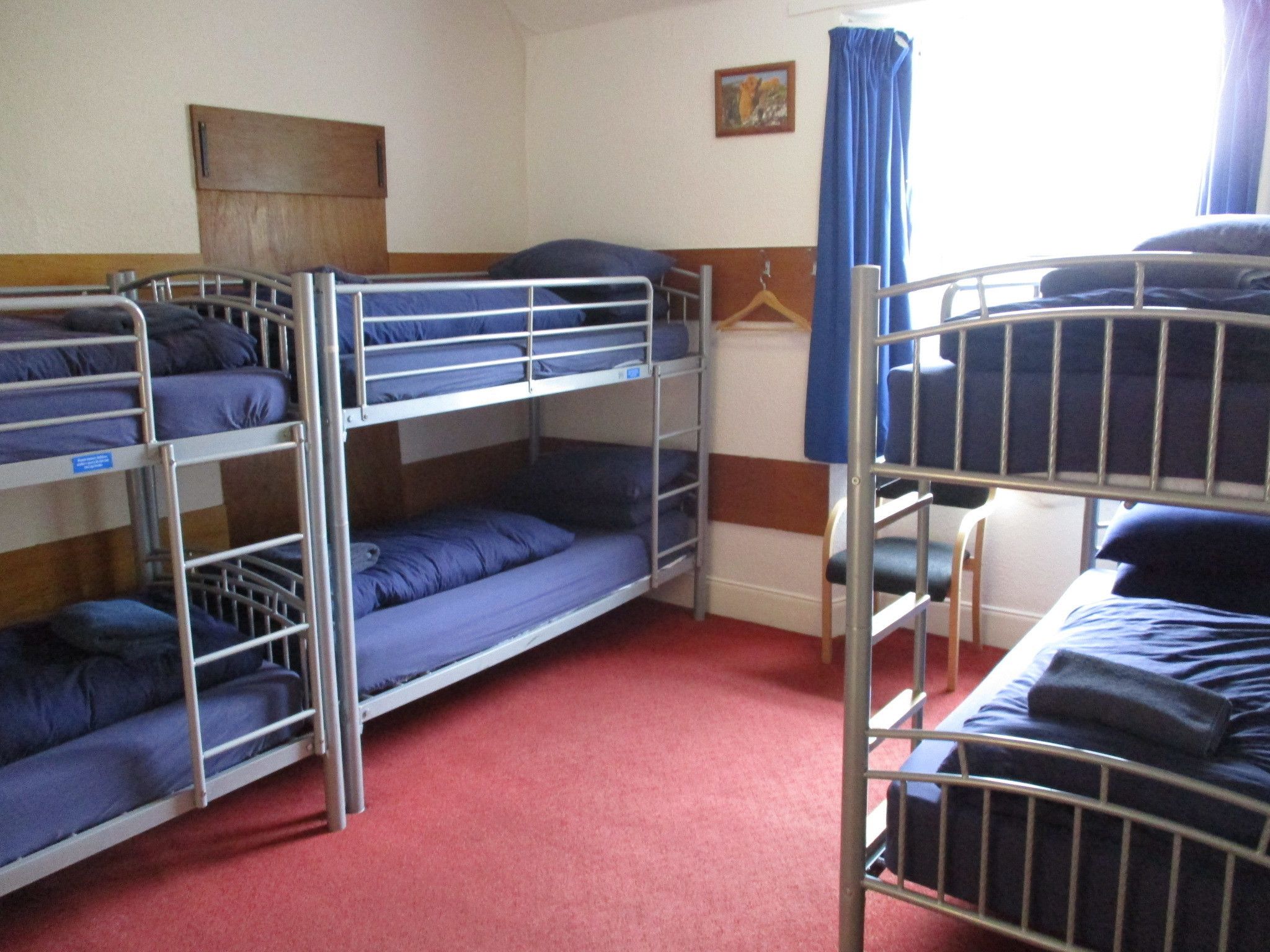 Habitación tipo dormitorio con literas, cabecero de madera, un cuadro en la pared y una cortina azul.