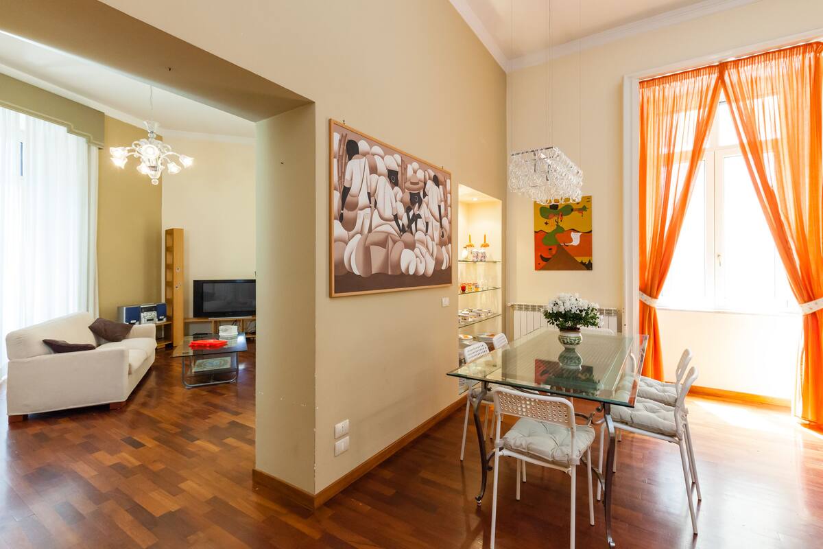 A modern living room with a glass dining table, white chairs, a sofa, and a TV.