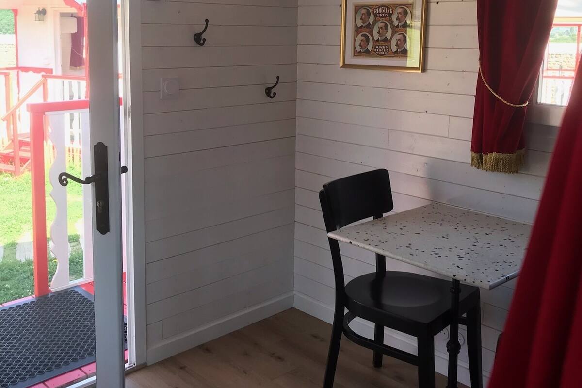 Habitació amb terra de fusta, una taula amb dues cadires, una porta amb maneta, un quadre a la paret i una cortina vermella.
