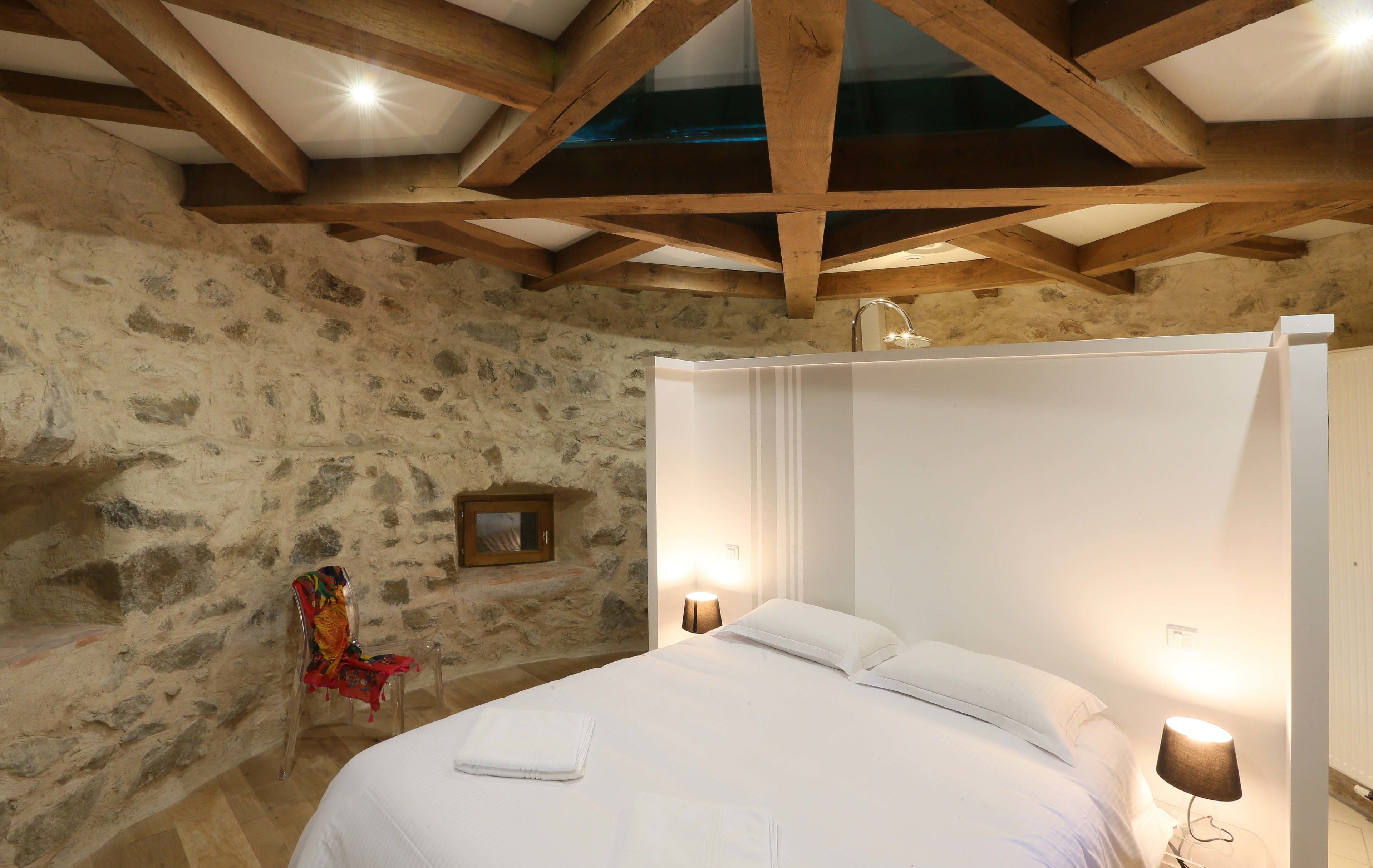 A bedroom with a stone wall, wooden beams, a bed with white linens, a chair, and two bedside lamps.