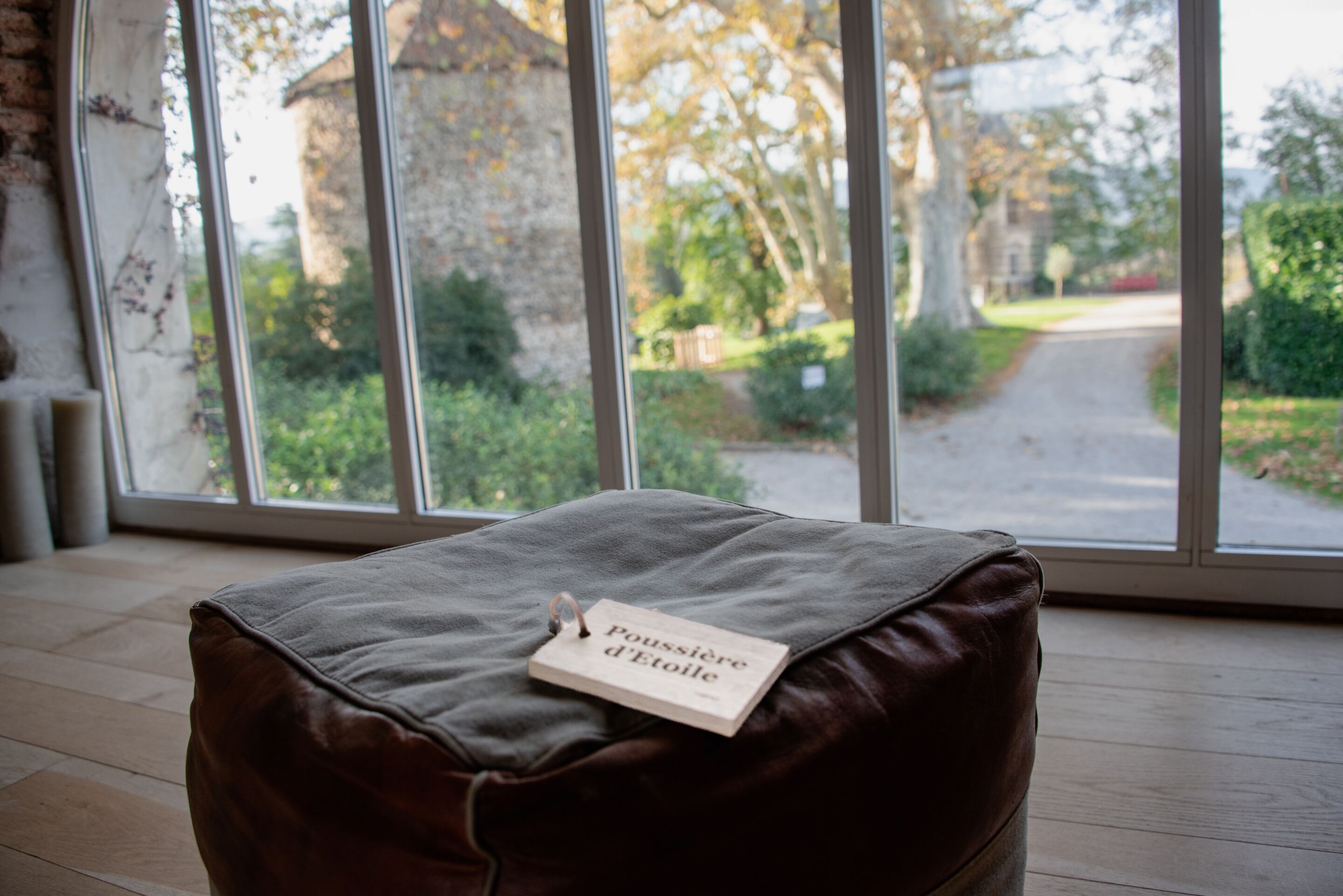 A brown leather ottoman with a tag reading "Poussée d'Etoile" in a room with wooden flooring and large windows overlooking a garden.