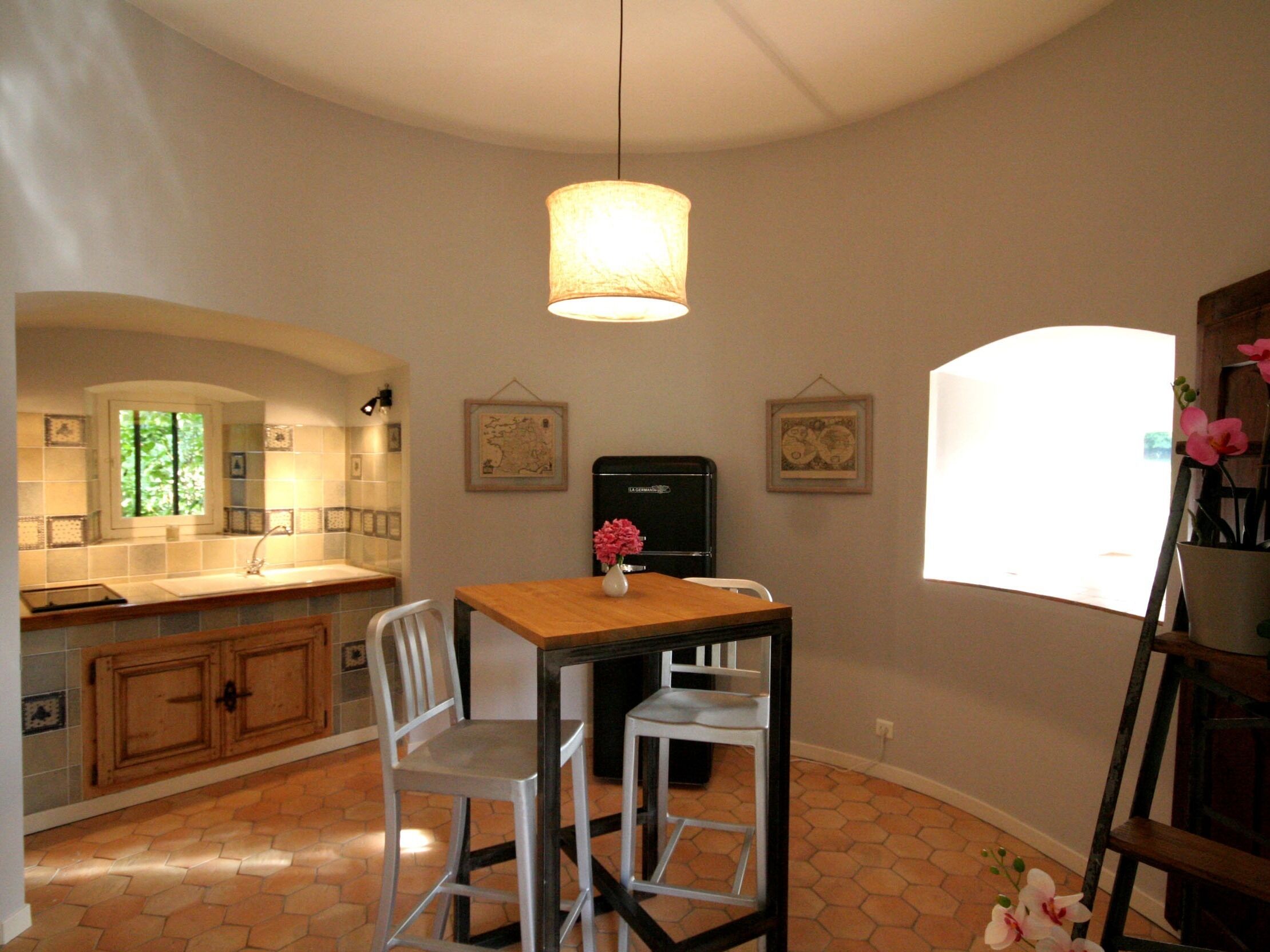 Una cocina con una mesa de madera, dos sillas blancas, una lámpara colgante, un refrigerador y una ventana con luz natural.