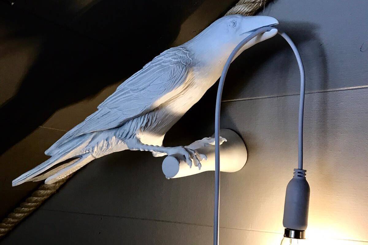 Escultura d'un ocell amb una llum penjada al bec, muntada a la paret.