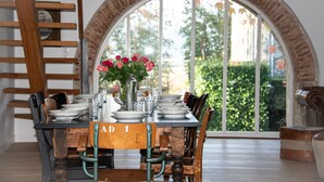 Een eettafel gedekt met borden, glazen en een bloevas, geplaatst voor een groot rondboogvenster met uitzicht op groen.
