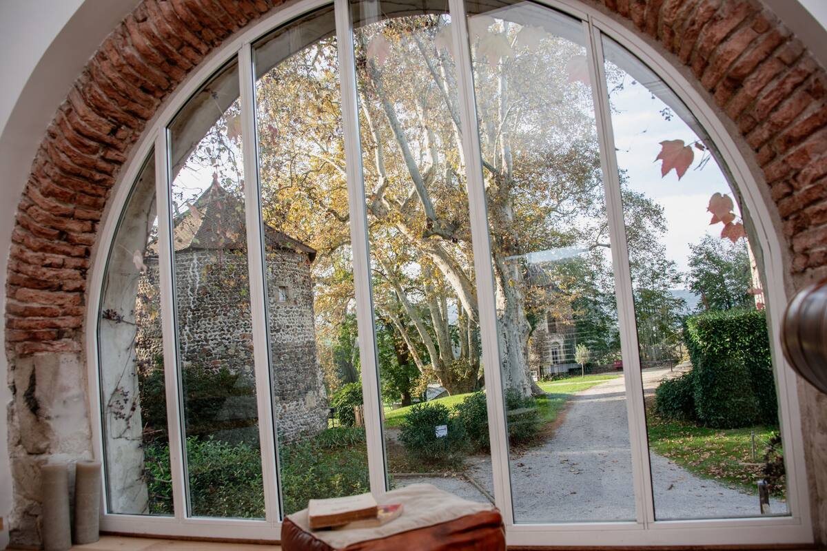 Vista a través d'una àmplia finestra d'arc amb arc de maó, cap a un jardí amb arbres i un camí.