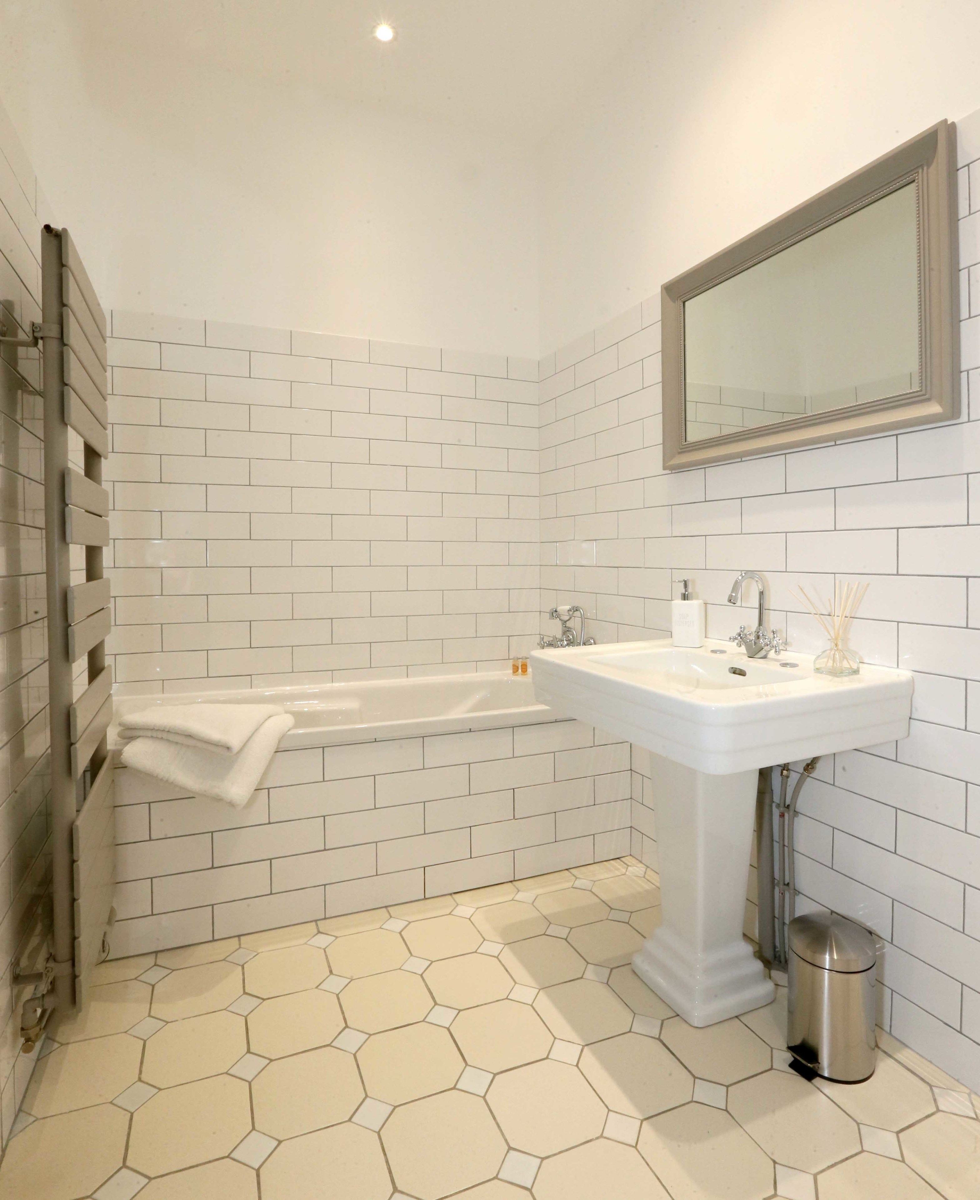 A bathroom with white hexagonal tiles, a white bathtub, a sink with a mirror above it, and a trash can.