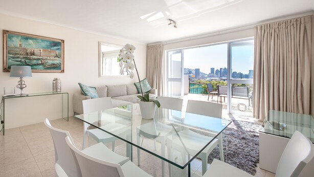Ein modernes Wohnzimmer mit einem Glastisch, weißen Sofas, Blick auf die Stadt durch große Fenster und einem Gemälde an der Wand.