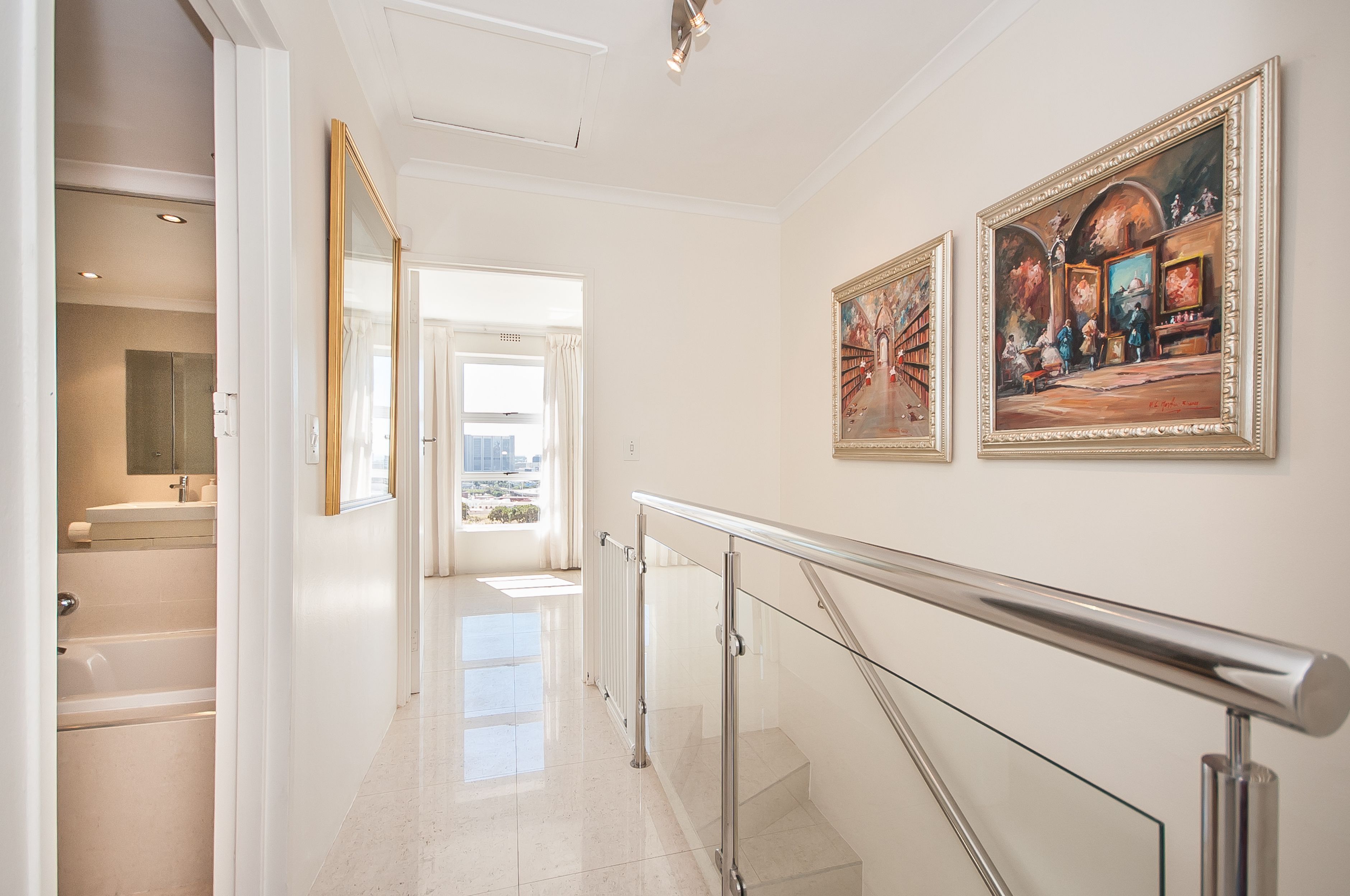 Een gang met een glazen balustrade, uitzicht naar buiten en twee ingelijste schilderijen aan de muur.