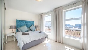 Ein modernes Schlafzimmer mit einem großen Bett, einem Nachttisch, einer Lampe und Blick auf eine Skyline durch große Fenster.