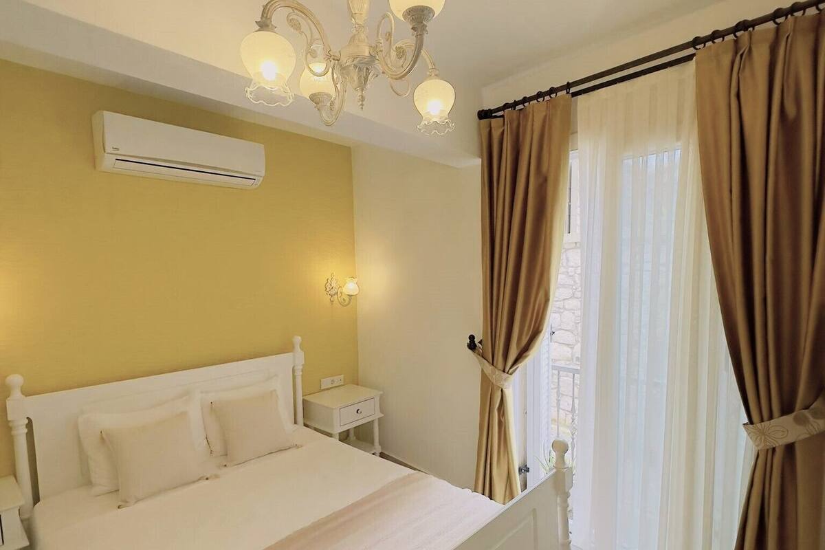 A bedroom with a bed, bedside tables, an air conditioning unit, a chandelier, and curtains.