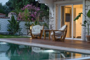 Un área junto a la piscina con dos sillones blancos acolchados y una pequeña mesa, rodeada de exuberante vegetación y plantas con flores.