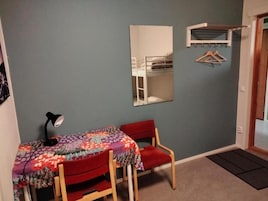 A small room with a single table and chair, a desk lamp, a mirror, and a coat rack.