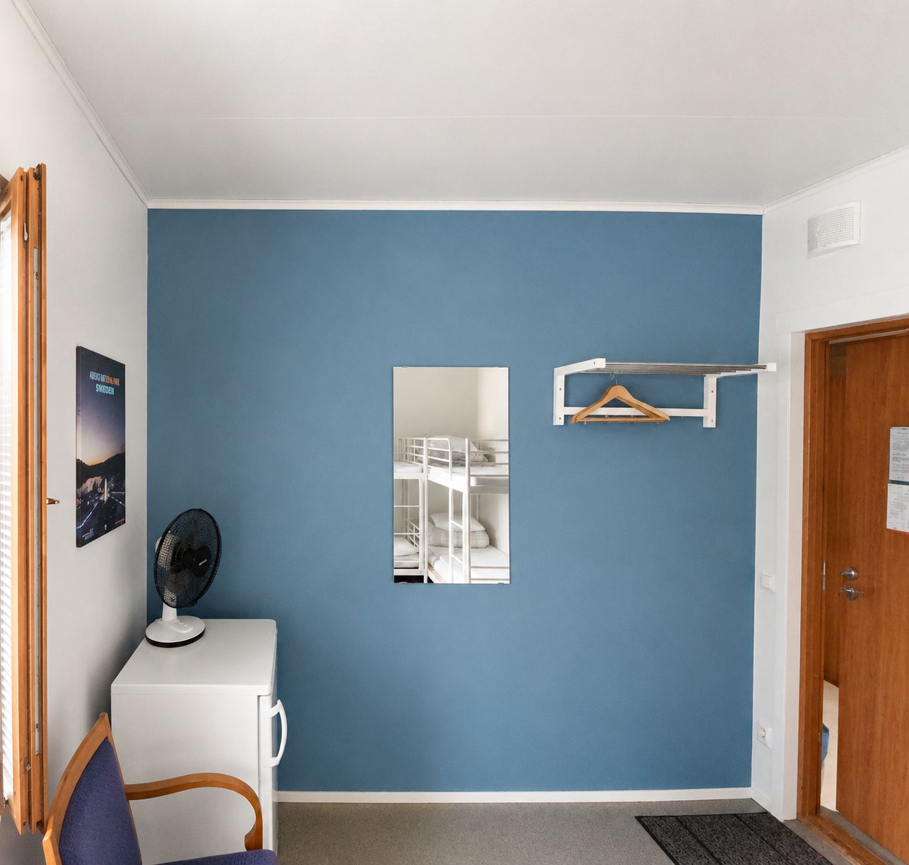 Une pièce avec un mur bleu, un bureau blanc avec un ventilateur, un miroir et une porte avec un cadre en bois.