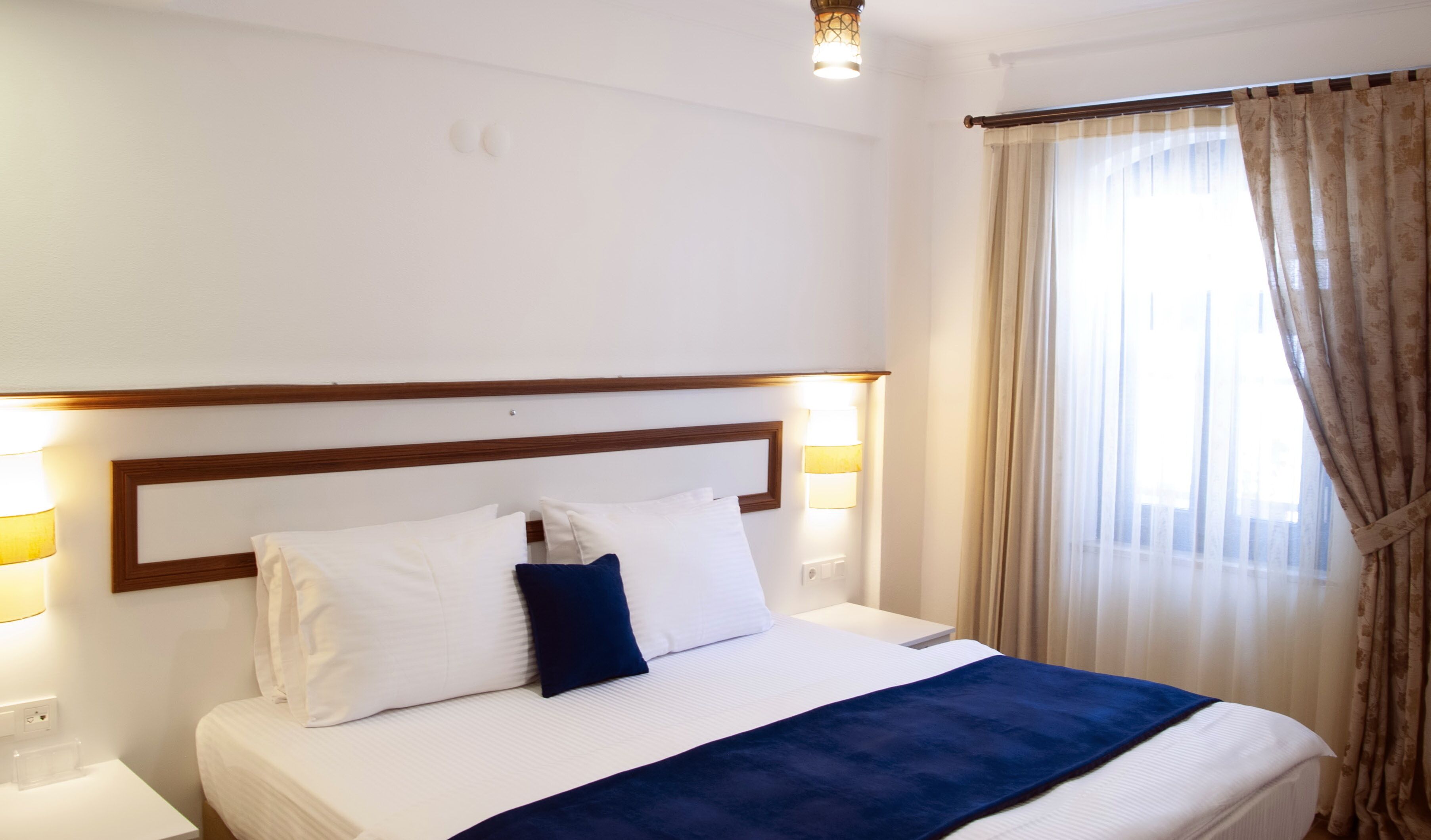 A hotel room with a large bed, a wooden headboard, two bedside lamps, and a window with curtains.
